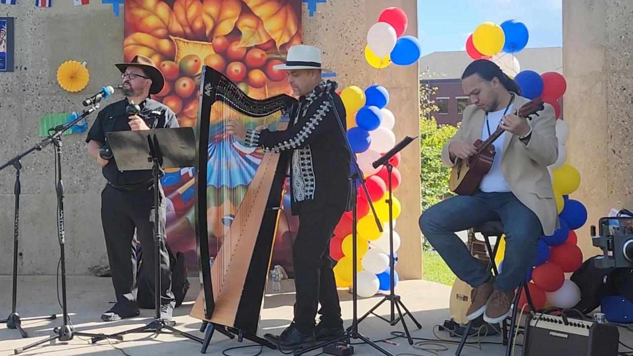 A trio plays music outdoors in front of a colorful mural and balloon arch; one plays a harp, one a guitar, one sings.