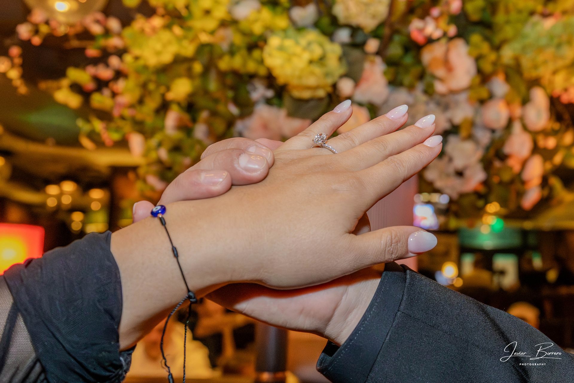 Two hands clasped together with rings, in front of a warm, floral-lit background.