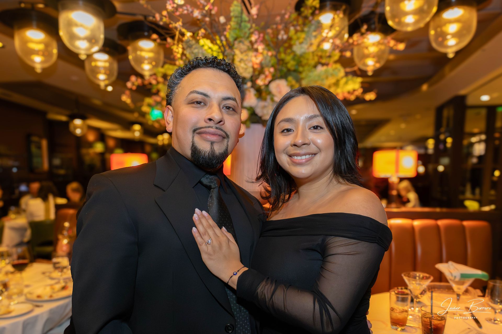 Couple posing in a warmly lit restaurant, with hanging globe lights and a set dining table in the background.