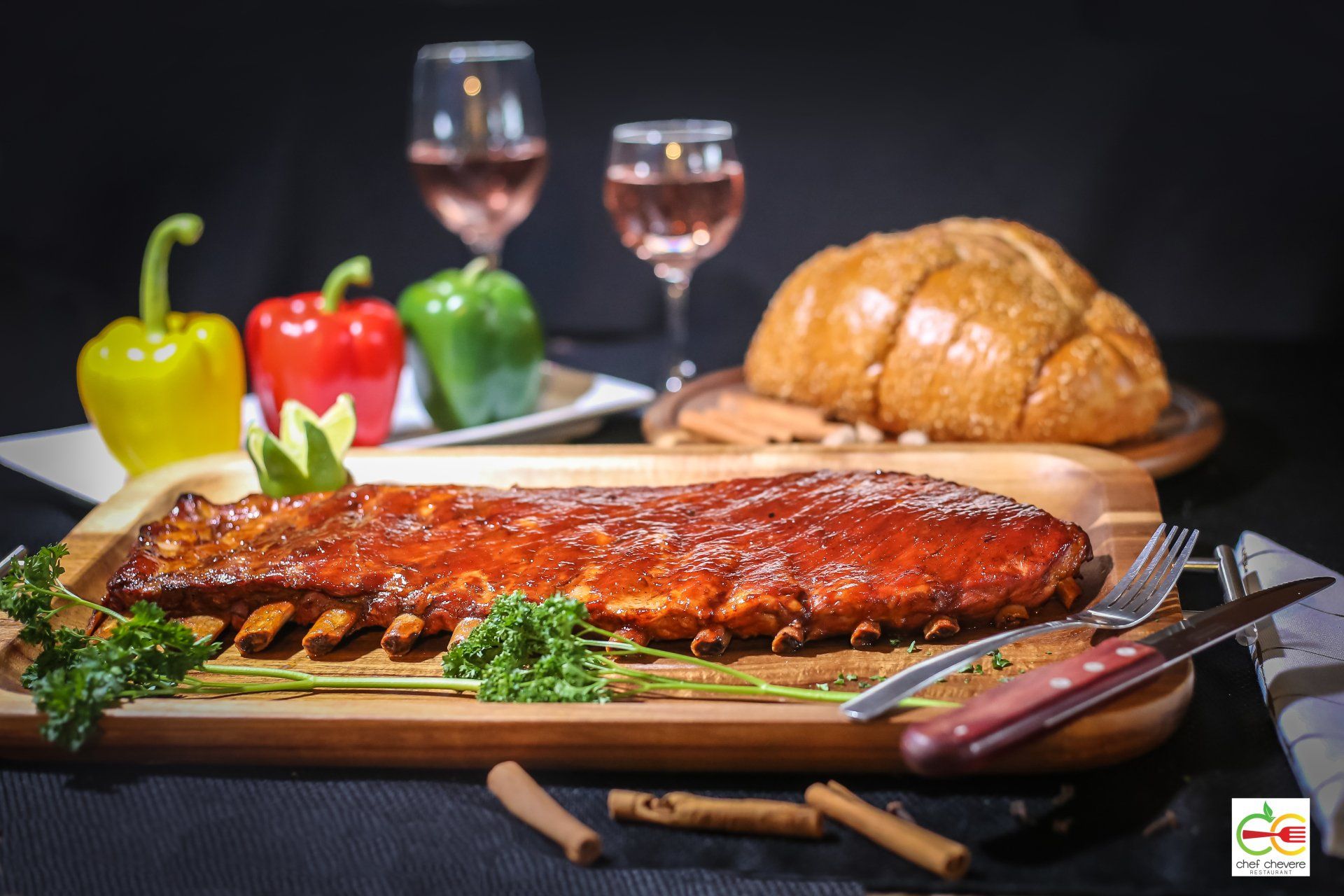 A rack of glazed ribs on a wooden platter with bell peppers, bread, and two wine glasses in the background.