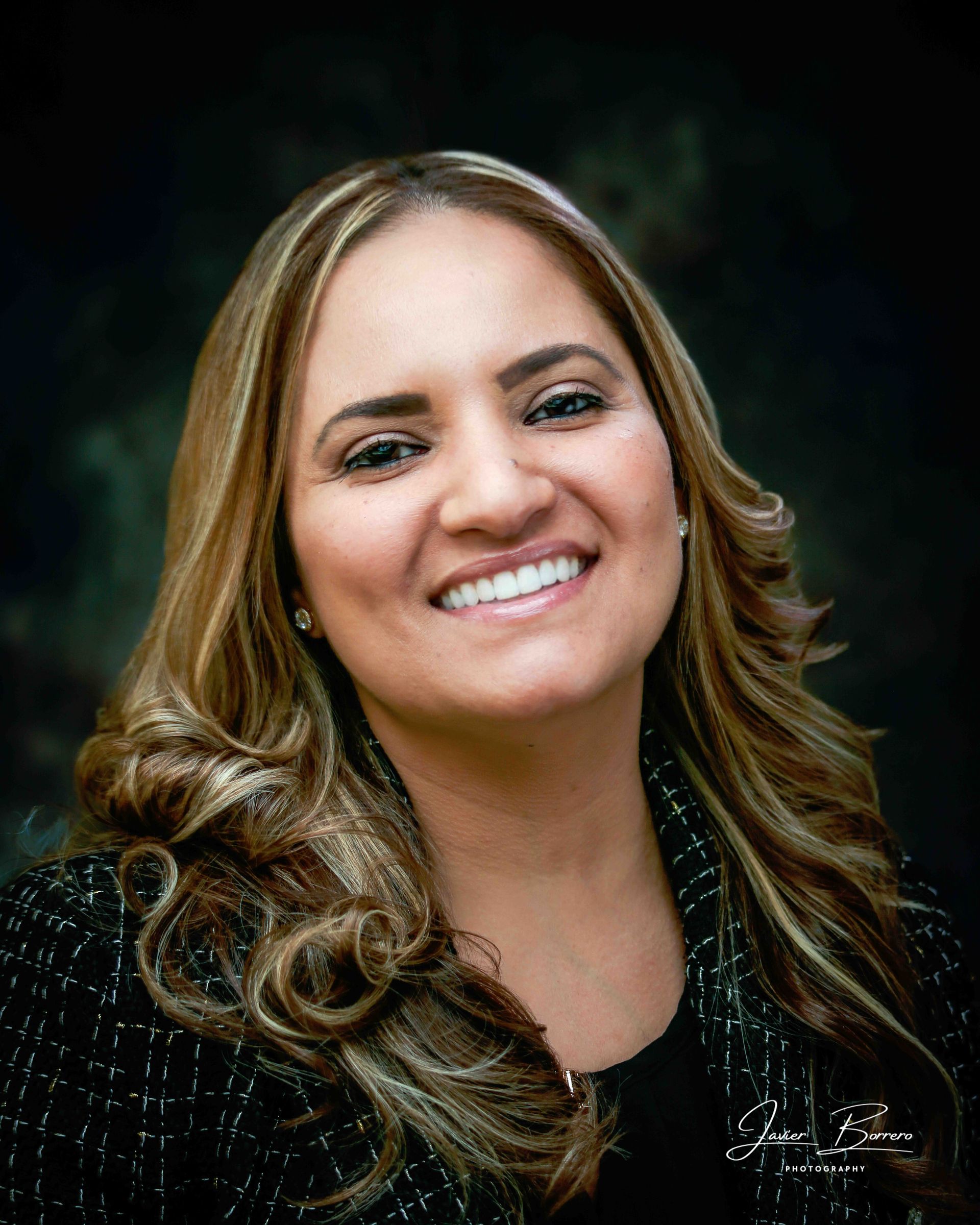 Smiling woman with long wavy hair in a dark patterned top against a black background