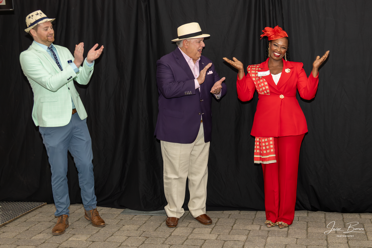 Three people onstage in colorful outfits and hats, clapping and smiling before a black curtain.