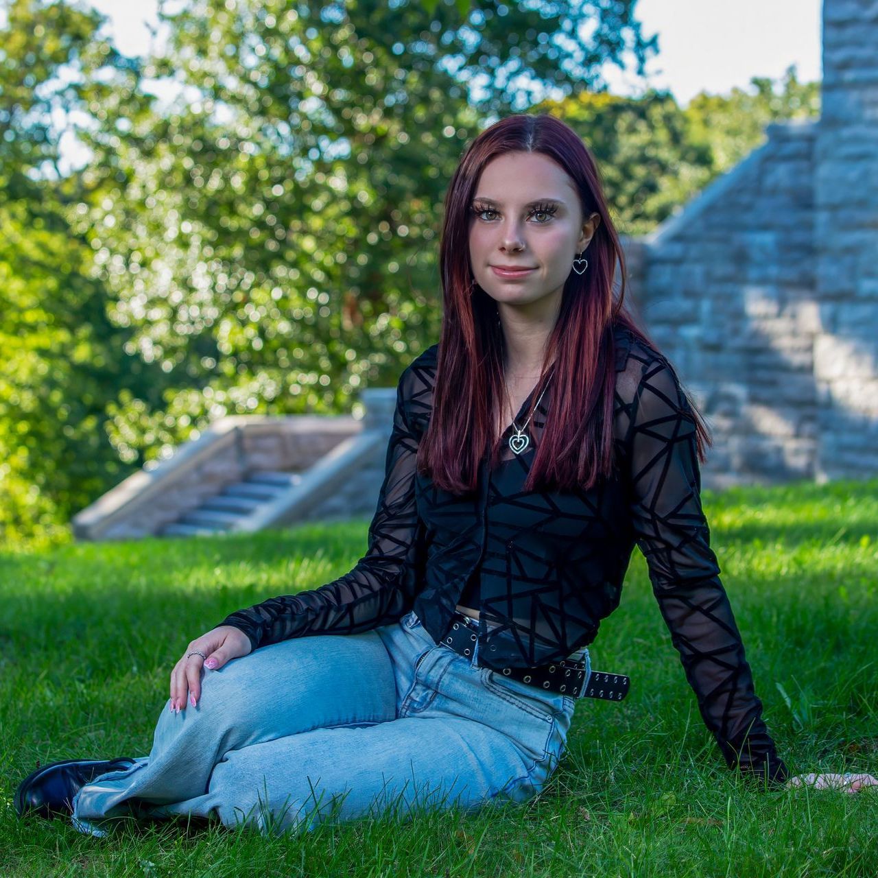 Person with long dark hair sitting on grass in a black top and light jeans near stone steps outdoors
