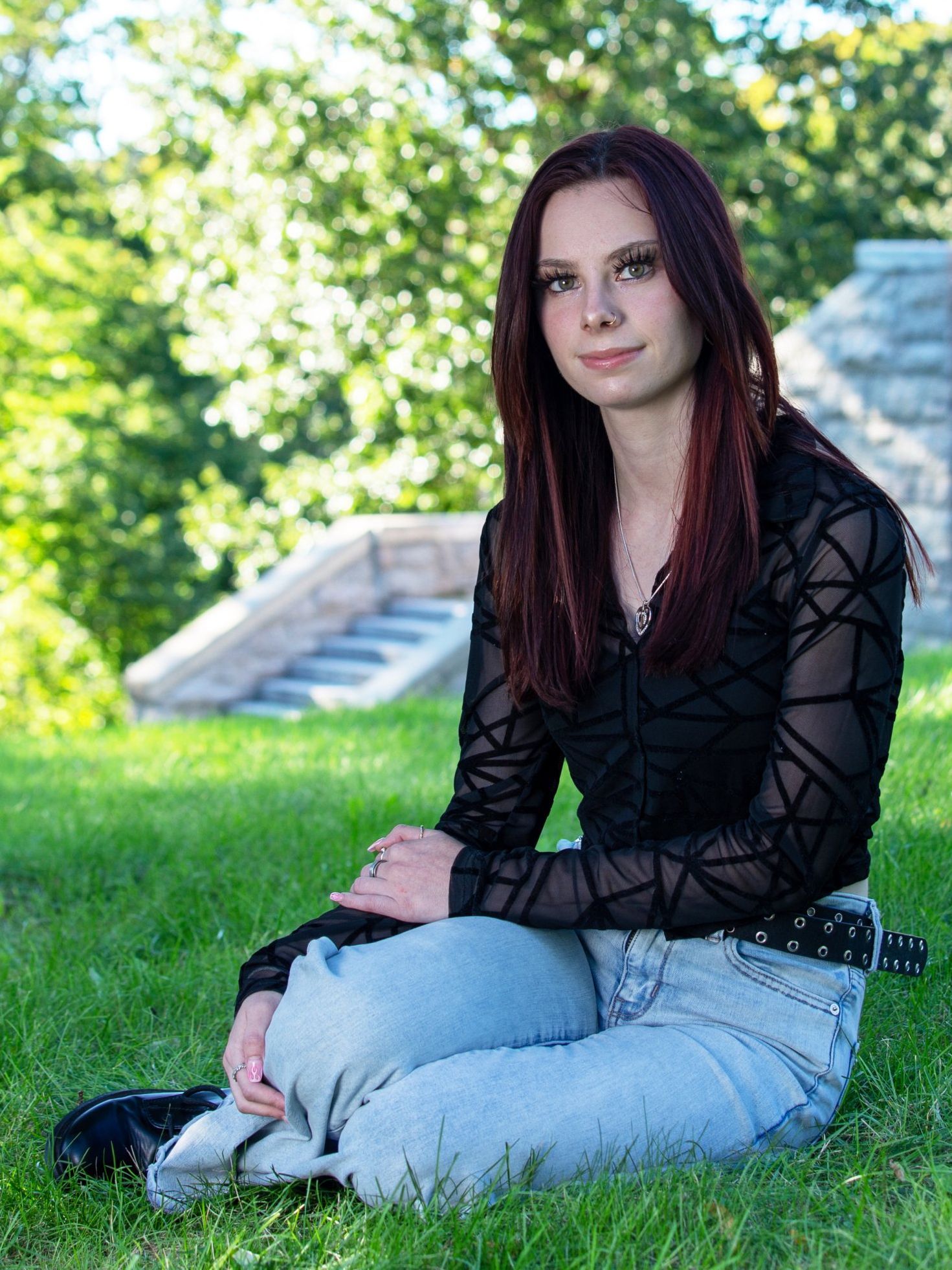 Woman sitting on grass outdoors in a black top and light jeans, with trees and steps in the background
