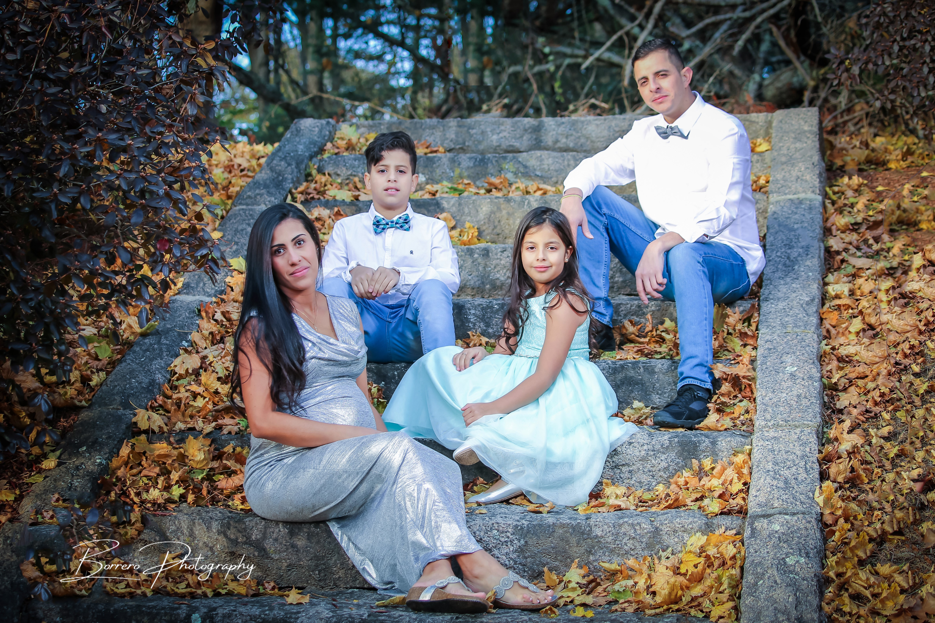 Family seated on stone steps outdoors, with autumn leaves and a man in white shirt on the top step.