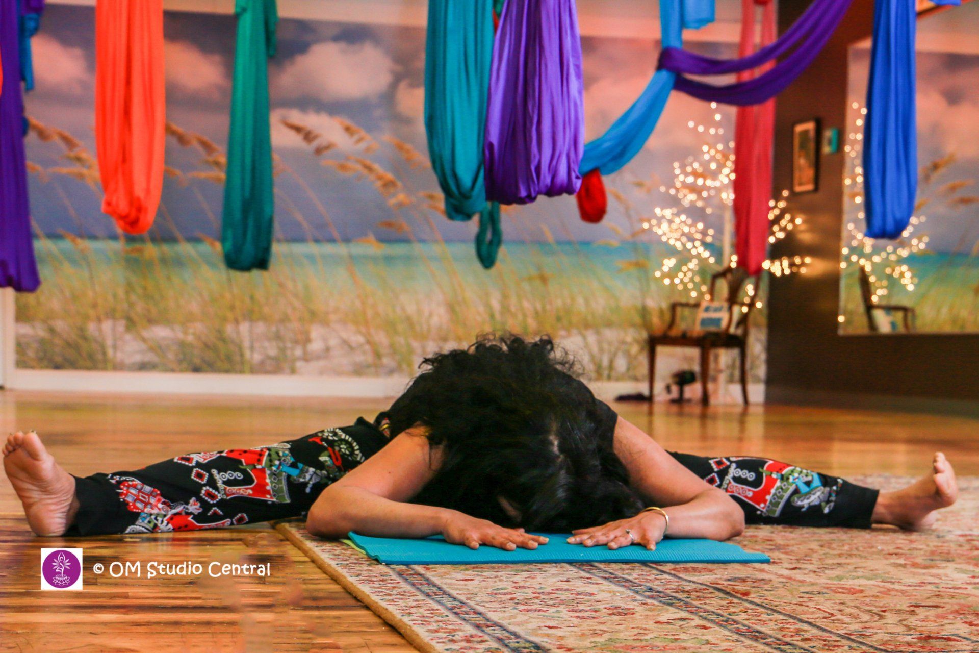 A person in colorful leggings performs a wide-legged forward fold on a mat in a studio with hanging aerial silks.