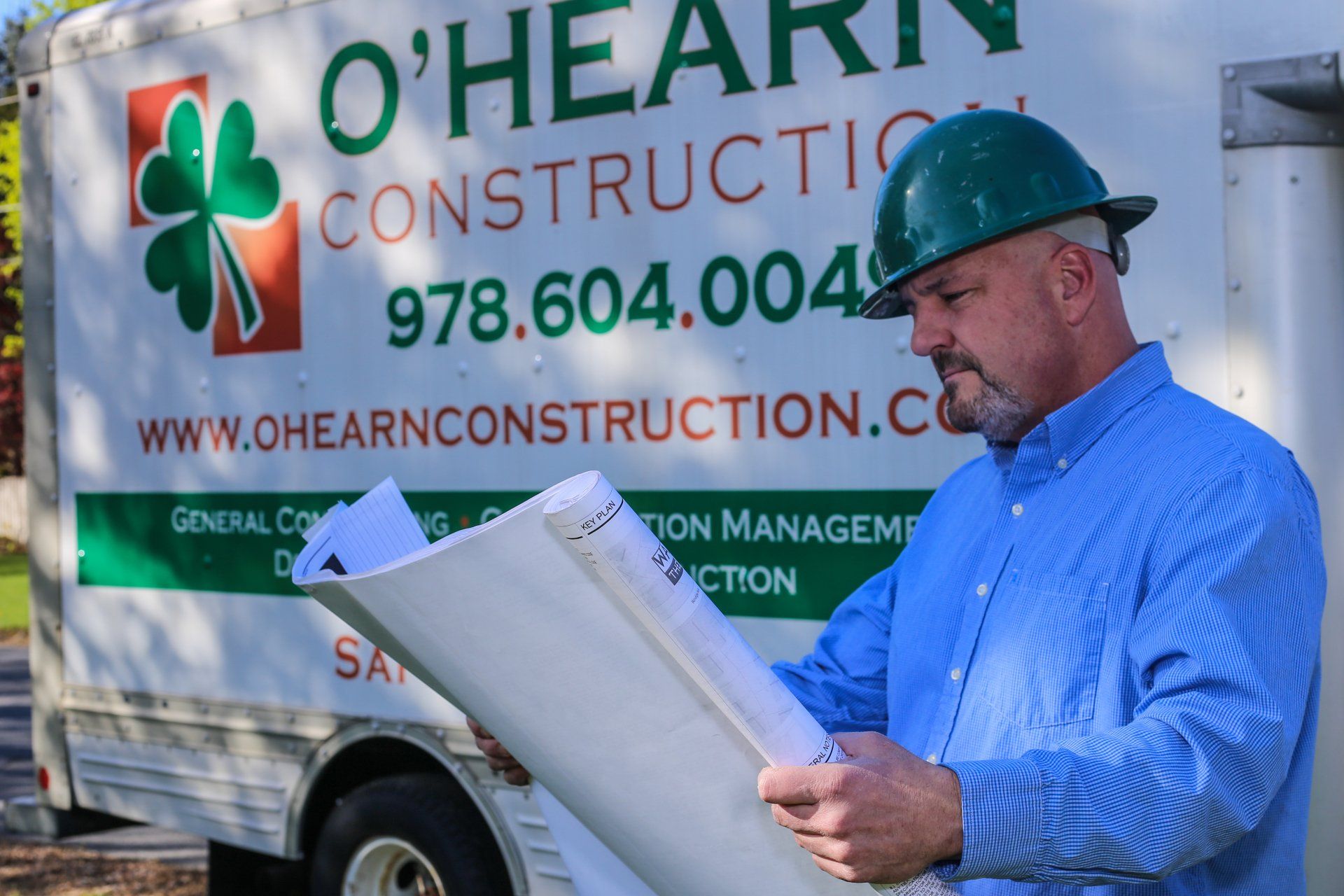A construction manager in a hard hat and blue shirt reviews blueprints in front of an O'Hearn Construction company truck.