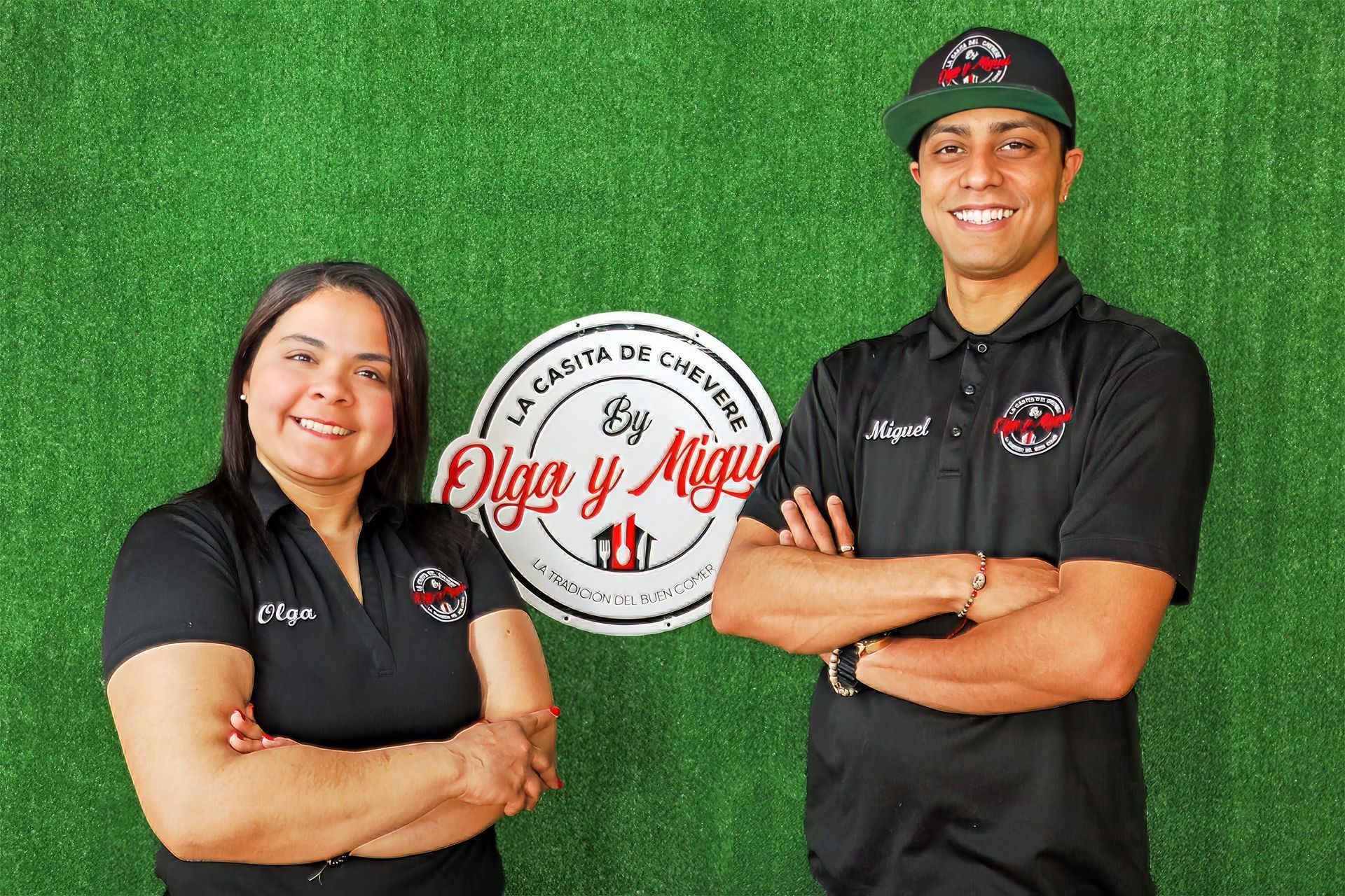 Olga and Miguel stand smiling with arms crossed in front of a green artificial grass wall with their restaurant logo.