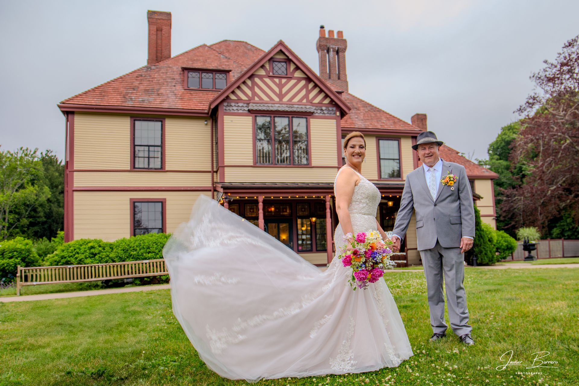 Bride and groom posing in front of a large house, with the bride’s white gown flowing and bouquet in hand.