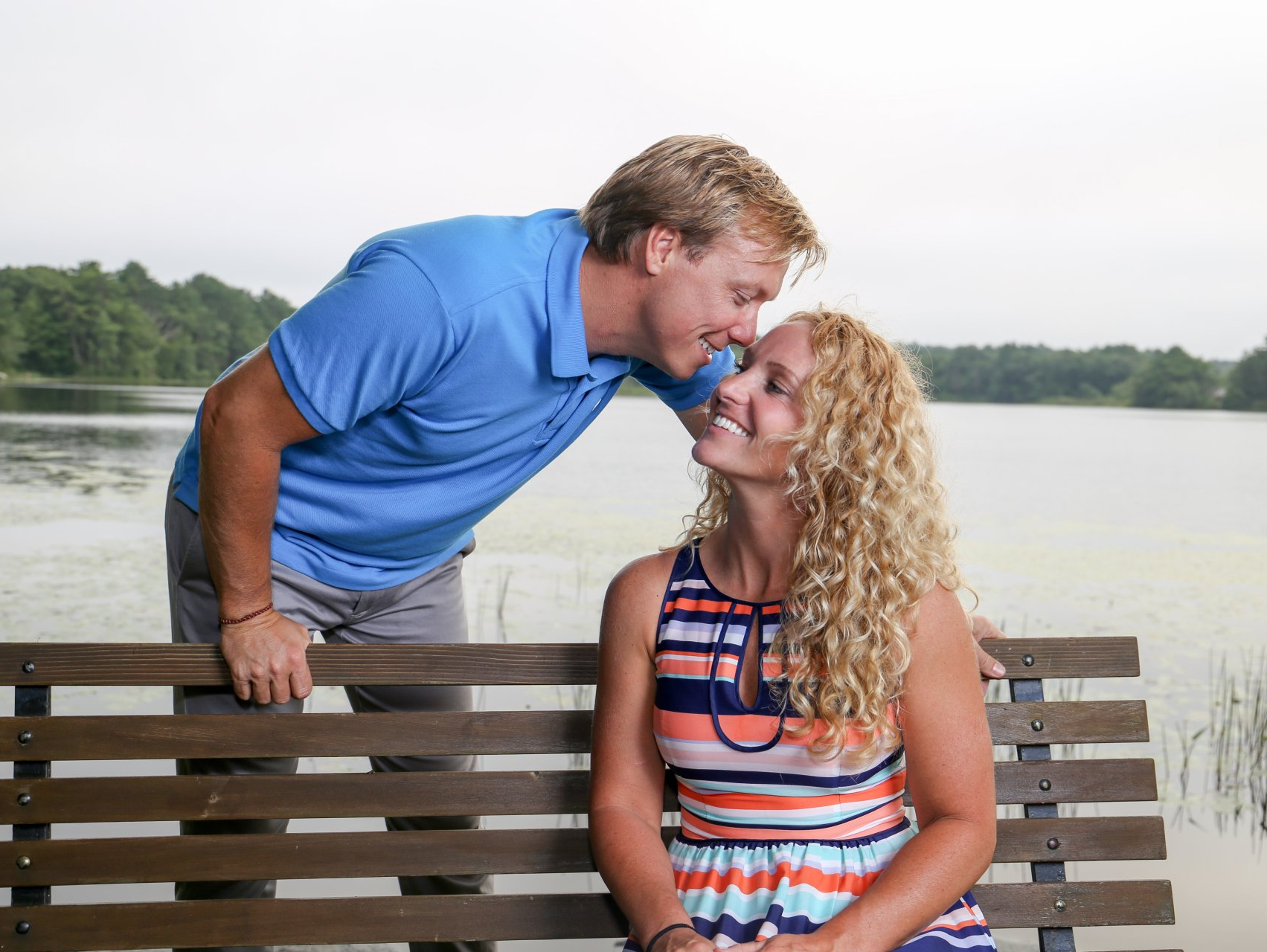 Couple sitting on a bench by the water, smiling and leaning in for a kiss.