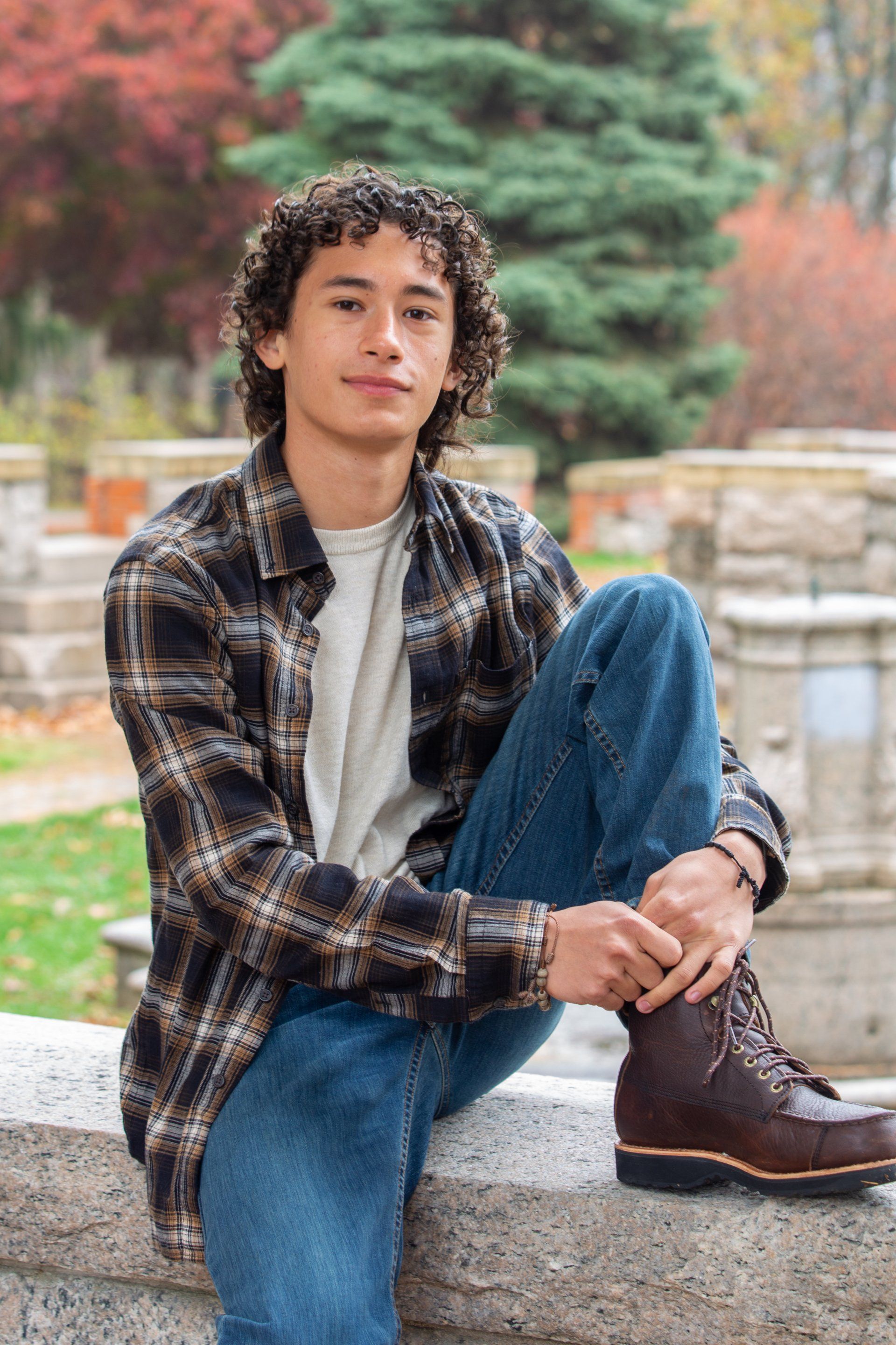 Young person in a plaid shirt and jeans sitting outdoors on a stone ledge with one leg crossed.