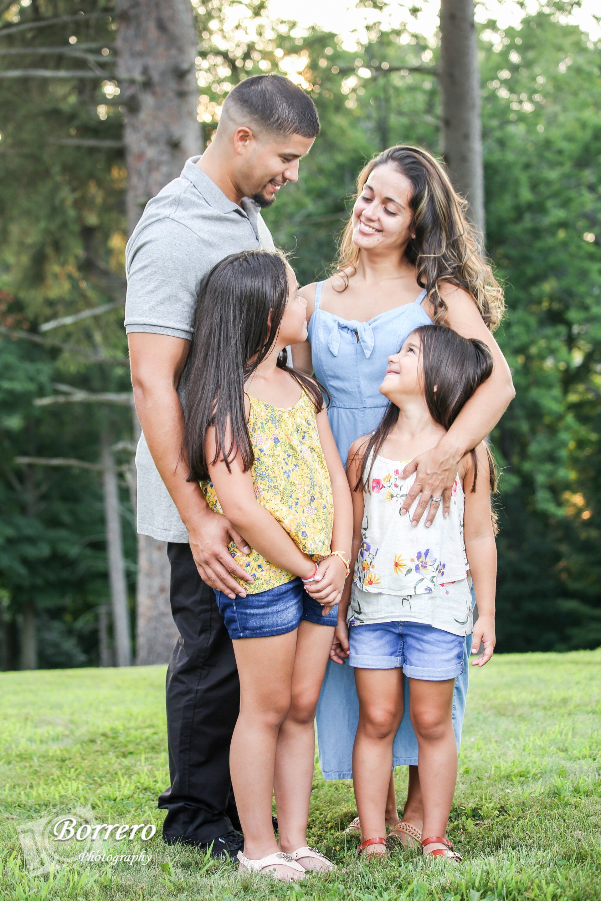 Family of four standing together on grass outdoors, smiling in a wooded park setting