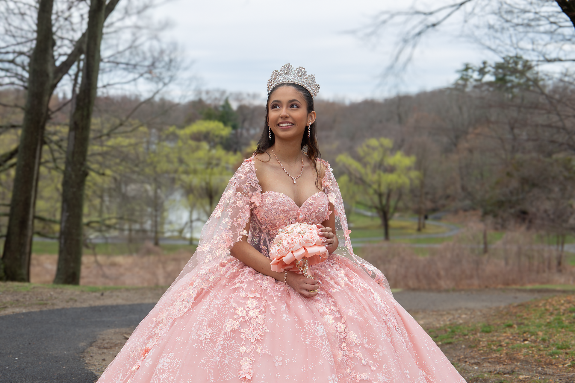Woman in a pink ball gown and tiara standing outdoors in a park, holding a bouquet.