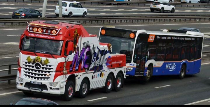 A brightly decorated tow truck towing a bus on a highway. The truck is red, white, and purple.