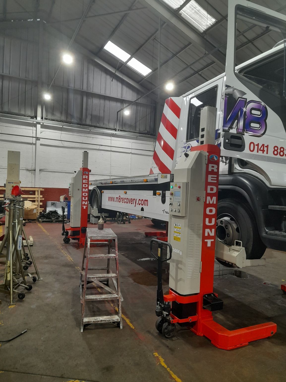 Red and white truck in a garage, lifted by two Redmount portable vehicle lifts. Step ladder stands nearby.