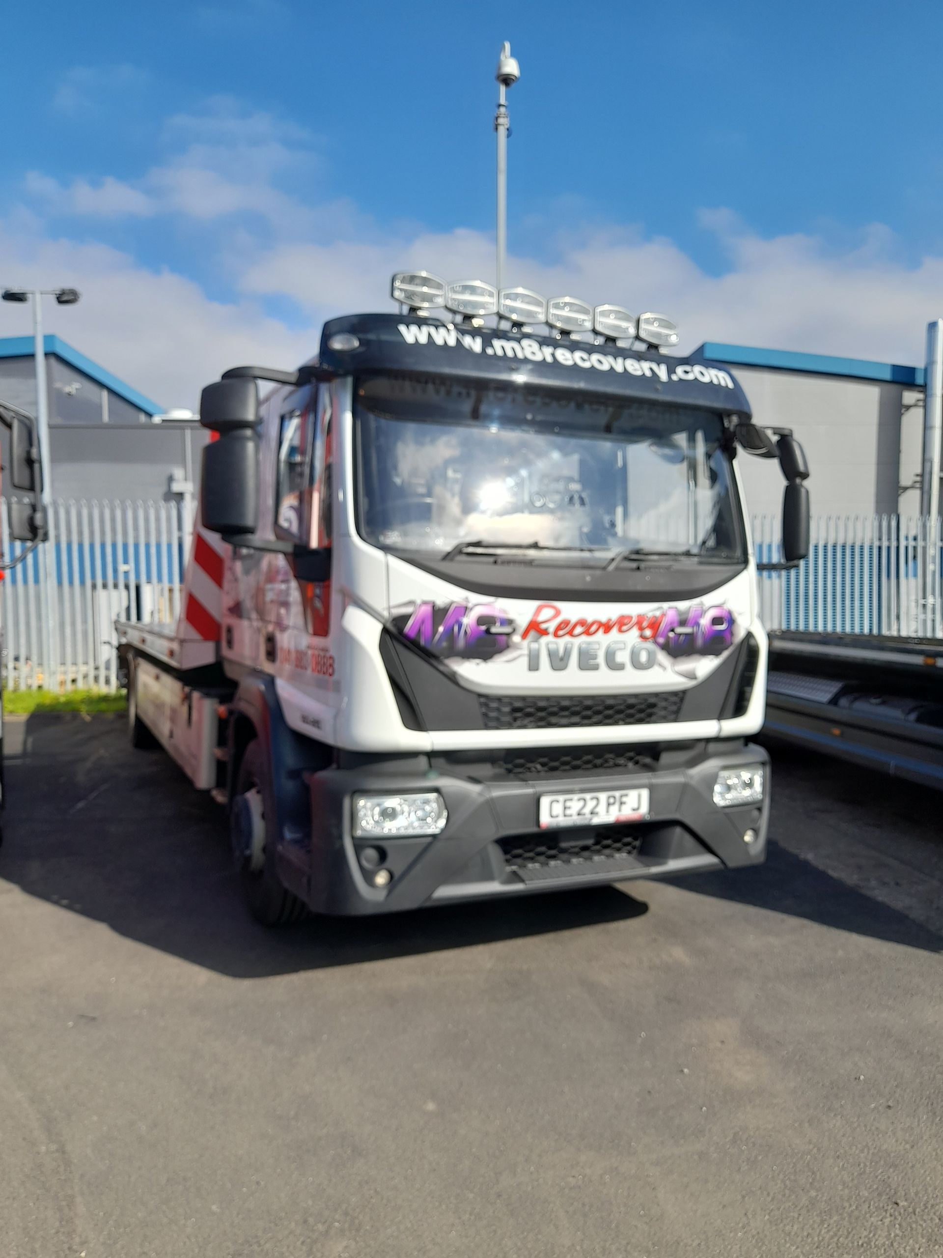 White Iveco tow truck with its flatbed in front of a blue sky and industrial buildings.