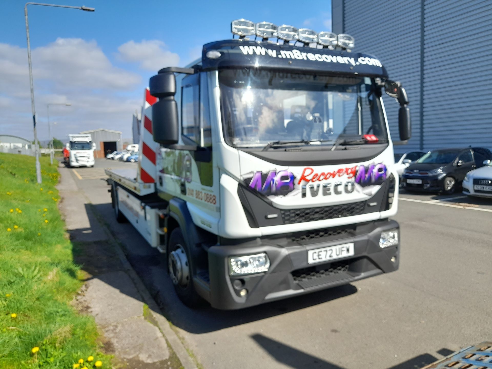 White Iveco tow truck with its flatbed in front of a blue sky and industrial buildings.