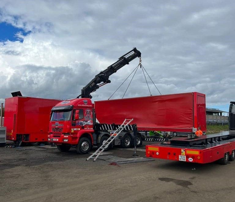 Red crane lifting a large red container, other red containers and truck visible. Cloudy sky.