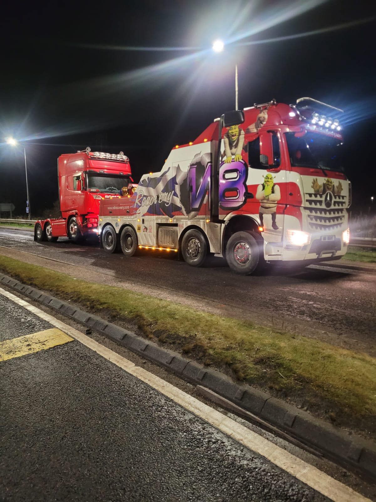 Red tow truck towing a red semi-truck at night. Bright headlights illuminate both vehicles.