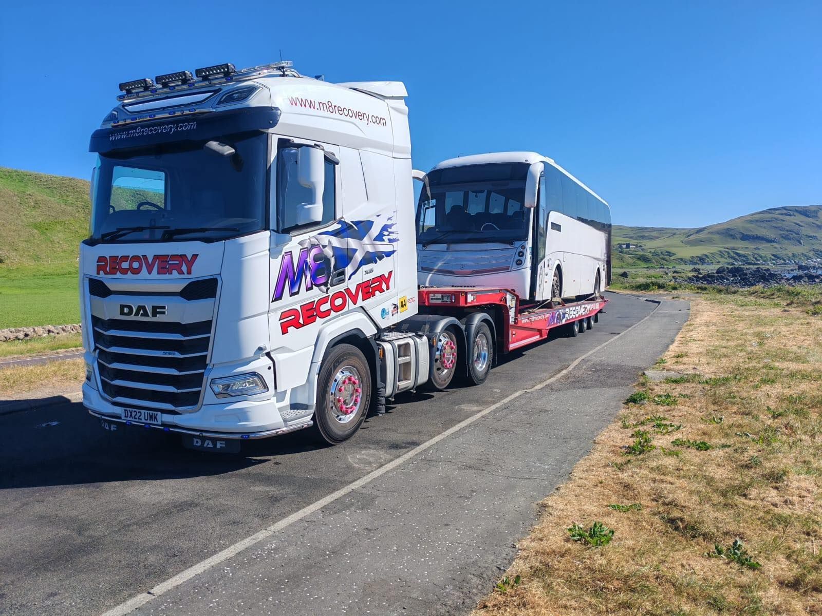 A white recovery truck with a bus on a trailer on a rural road under a blue sky.
