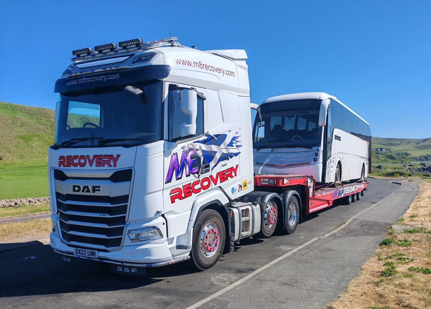 White recovery truck towing a white bus on a rural road under a blue sky. 