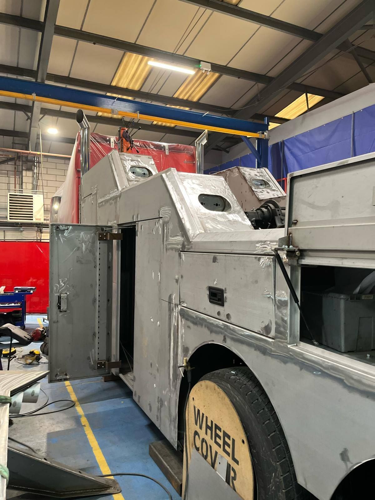 A large, gray vehicle being worked on in a workshop. Open door reveals interior details, with wheel cover visible.