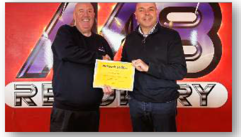 Two men in front of a red sign, one presenting a yellow certificate.