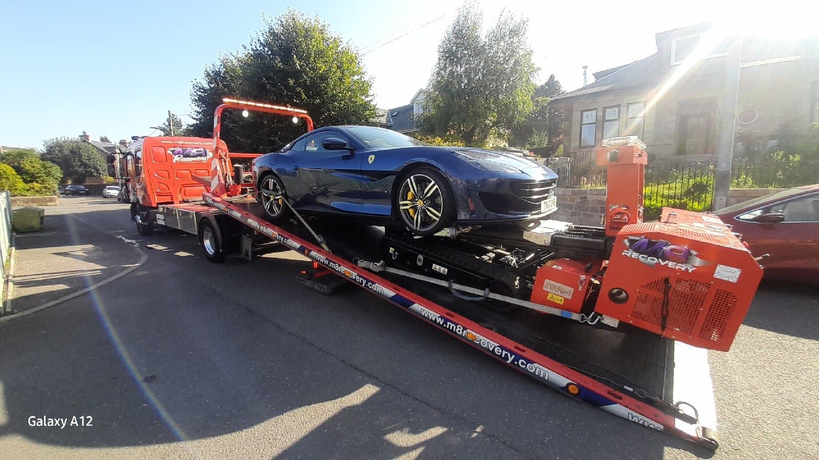 Blue Ferrari being loaded onto a flatbed tow truck on a sunny street.