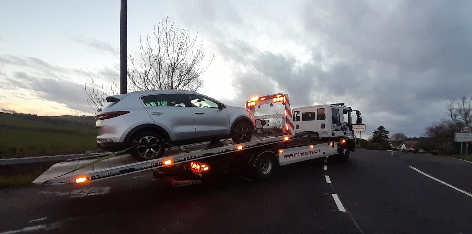 A white SUV is loaded on a tow truck on a road under a cloudy sky.