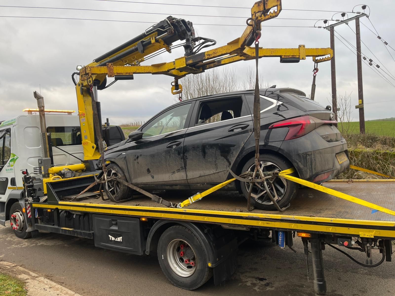 A black car being towed by a yellow tow truck on a road; cloudy day.