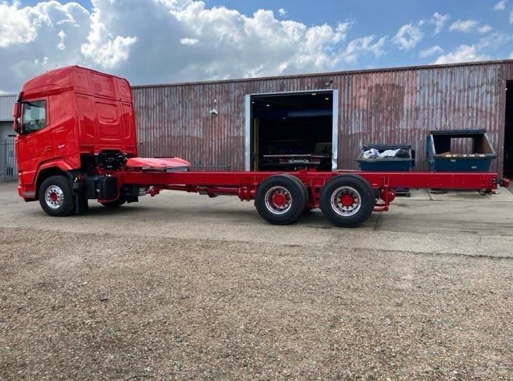 Red truck chassis and cab against a weathered building under a cloudy sky.