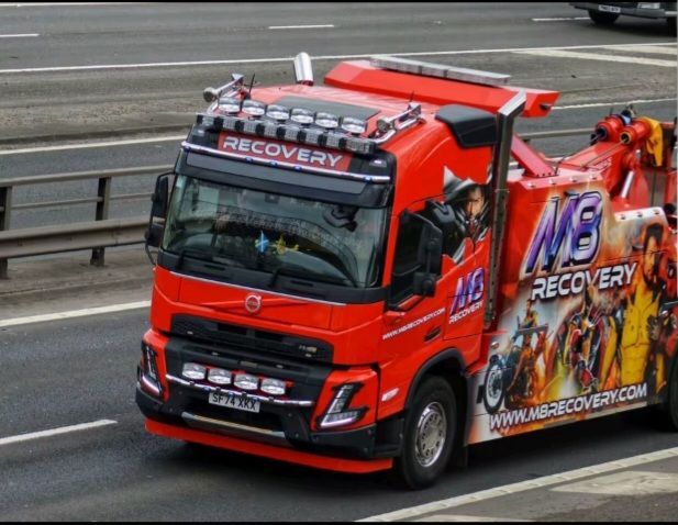 Red recovery tow truck driving on a highway, with bold white lettering and a damaged car on the flatbed.