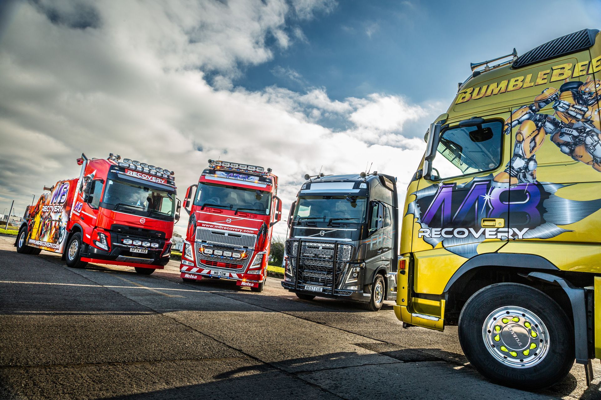 Four brightly colored recovery trucks parked on a road under a cloudy sky. The truck in the foreground is yellow.