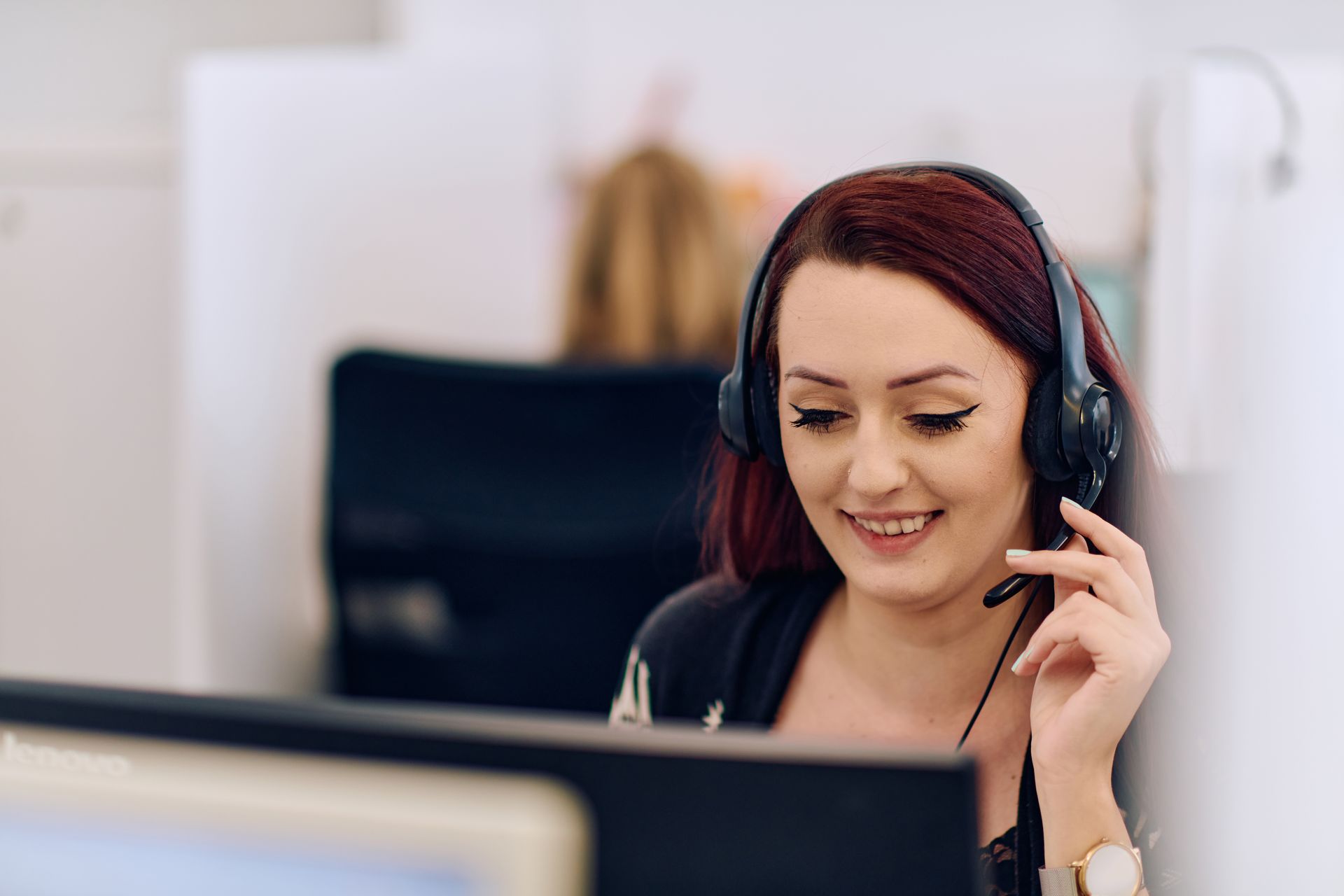 Woman with headset smiles while working at a computer in an office setting.