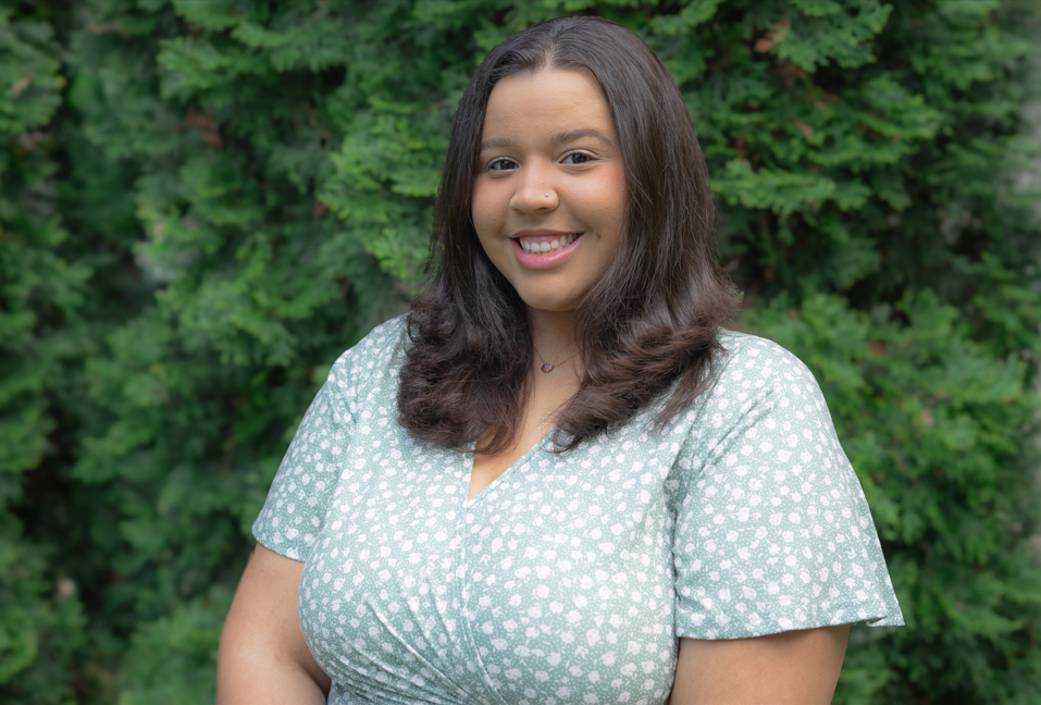 Smiling woman in a light green patterned dress standing outdoors in front of green foliage