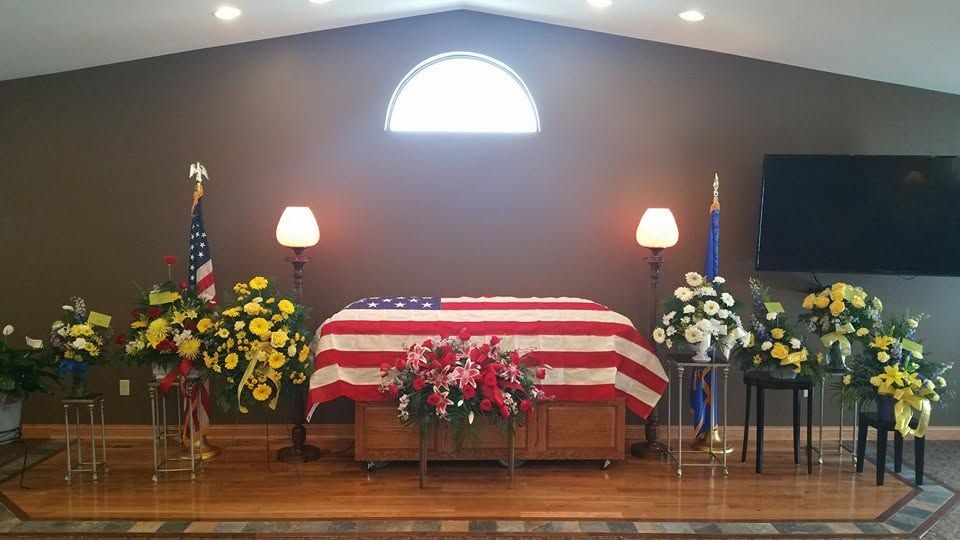 Funeral casket draped in an American flag, surrounded by floral arrangements on a church stage.