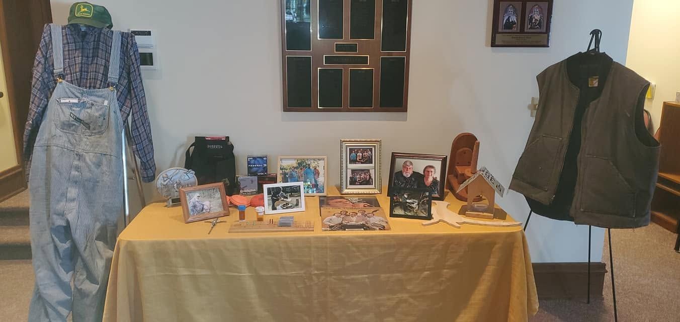 Display table with framed photos, books, and crafts, flanked by a patterned dress and a green vest on stands