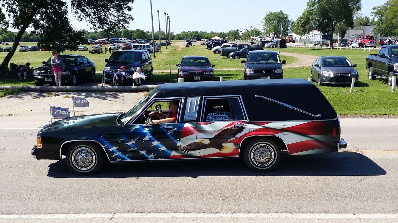 Black hearse with American flag paint parked by roadside at a car show