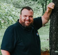 Smiling man in a black polo leaning against a tree in a green outdoor setting