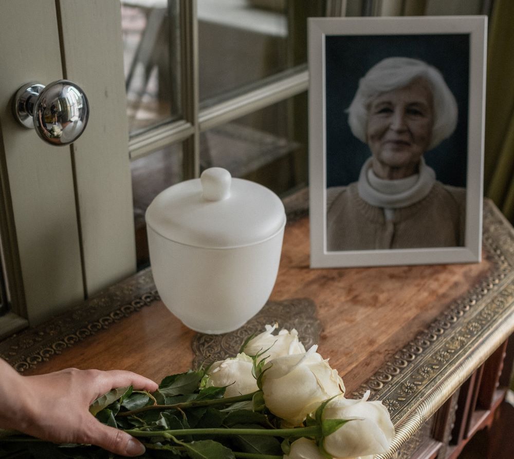 White roses placed beside a framed memorial portrait and a white urn on a wooden table