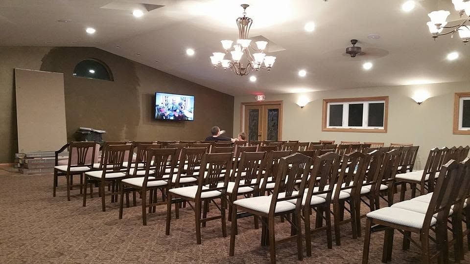 Rows of brown chairs in a banquet hall with chandeliers, a TV on the wall, and carpeted floor