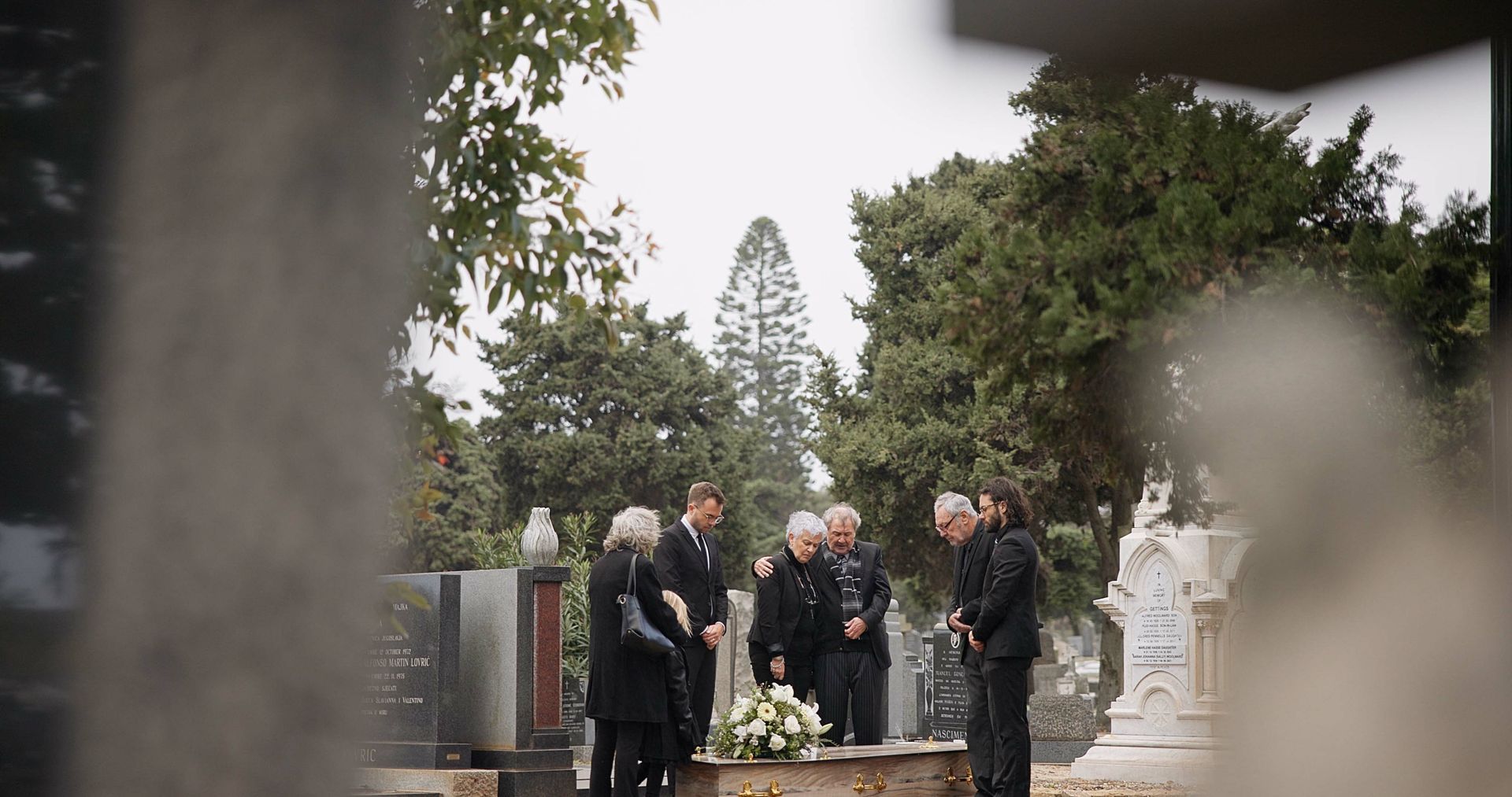 Mourners gathered at a cemetery around a flower-covered grave for a funeral ceremony.