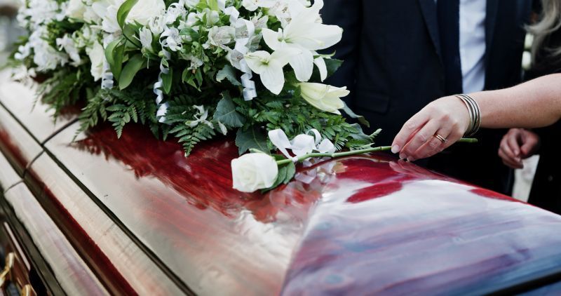 White flowers on a glossy red coffin as a hand adjusts the bouquet at a funeral