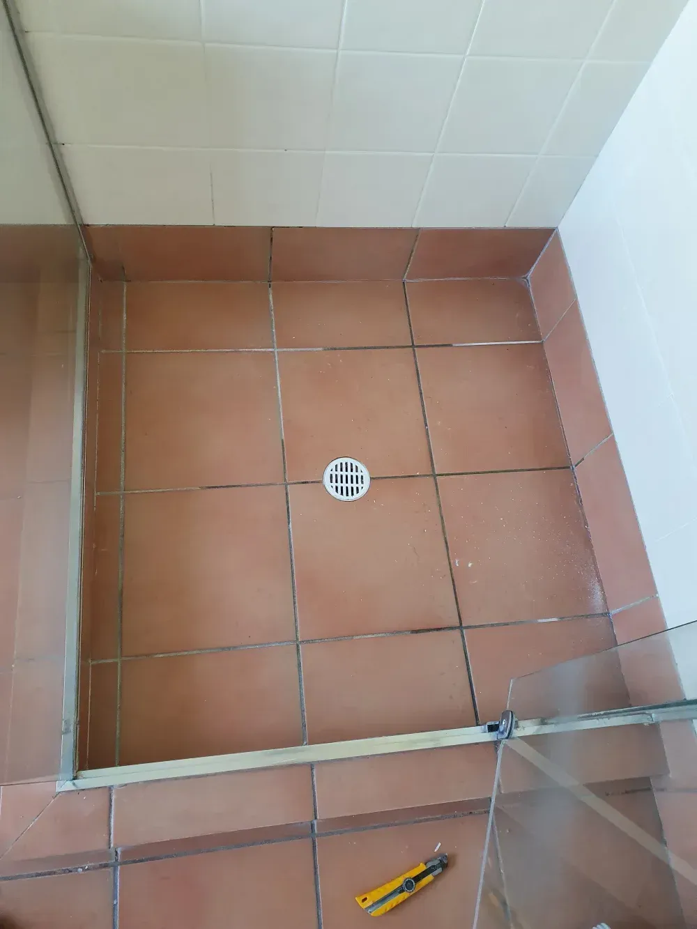 Small tiled shower floor with a center drain and a yellow utility knife near the bottom edge.