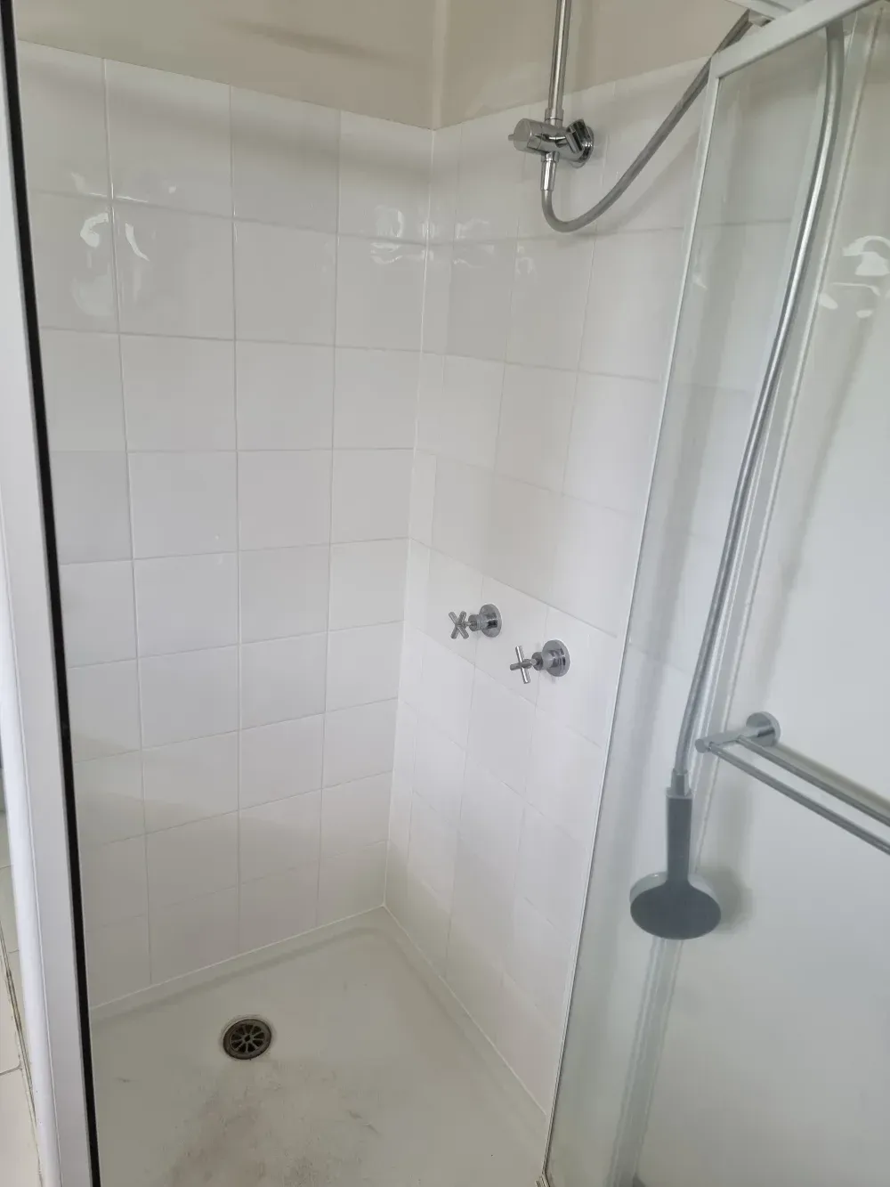 Empty white tiled shower with a drain and glass shower door.
