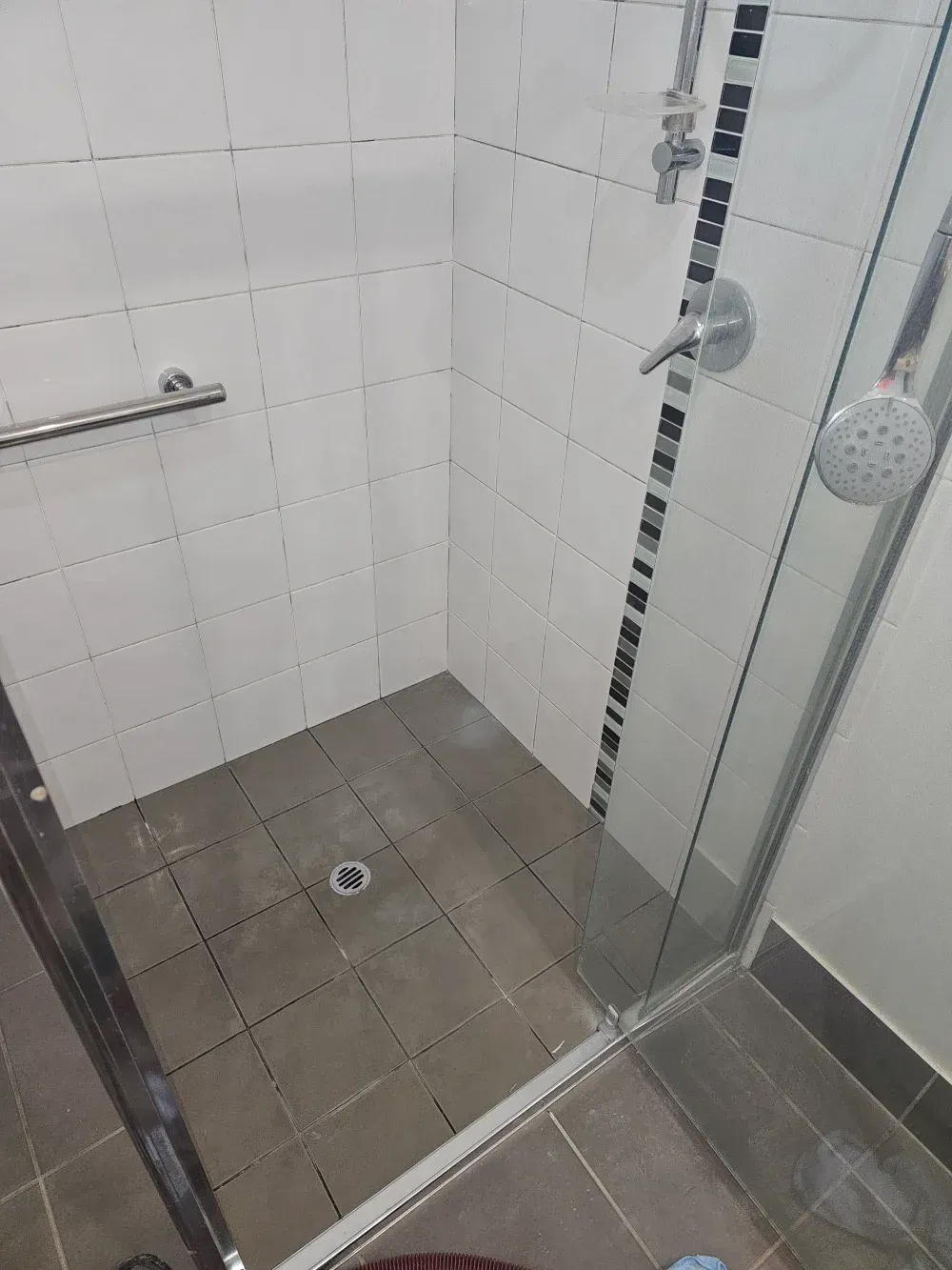 Small tiled shower corner with glass door, gray floor tiles, wall grab bar, and drain.