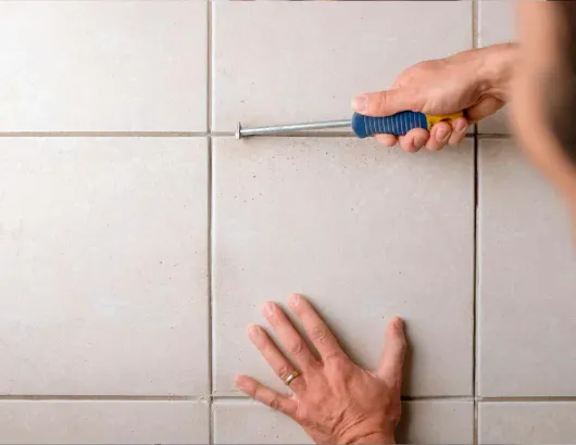 Hands using a screwdriver to pry a tile from a tiled wall
