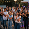 Público aplaudiendo en un evento nocturno al aire libre con guirnaldas de luces y un escenario al fondo.