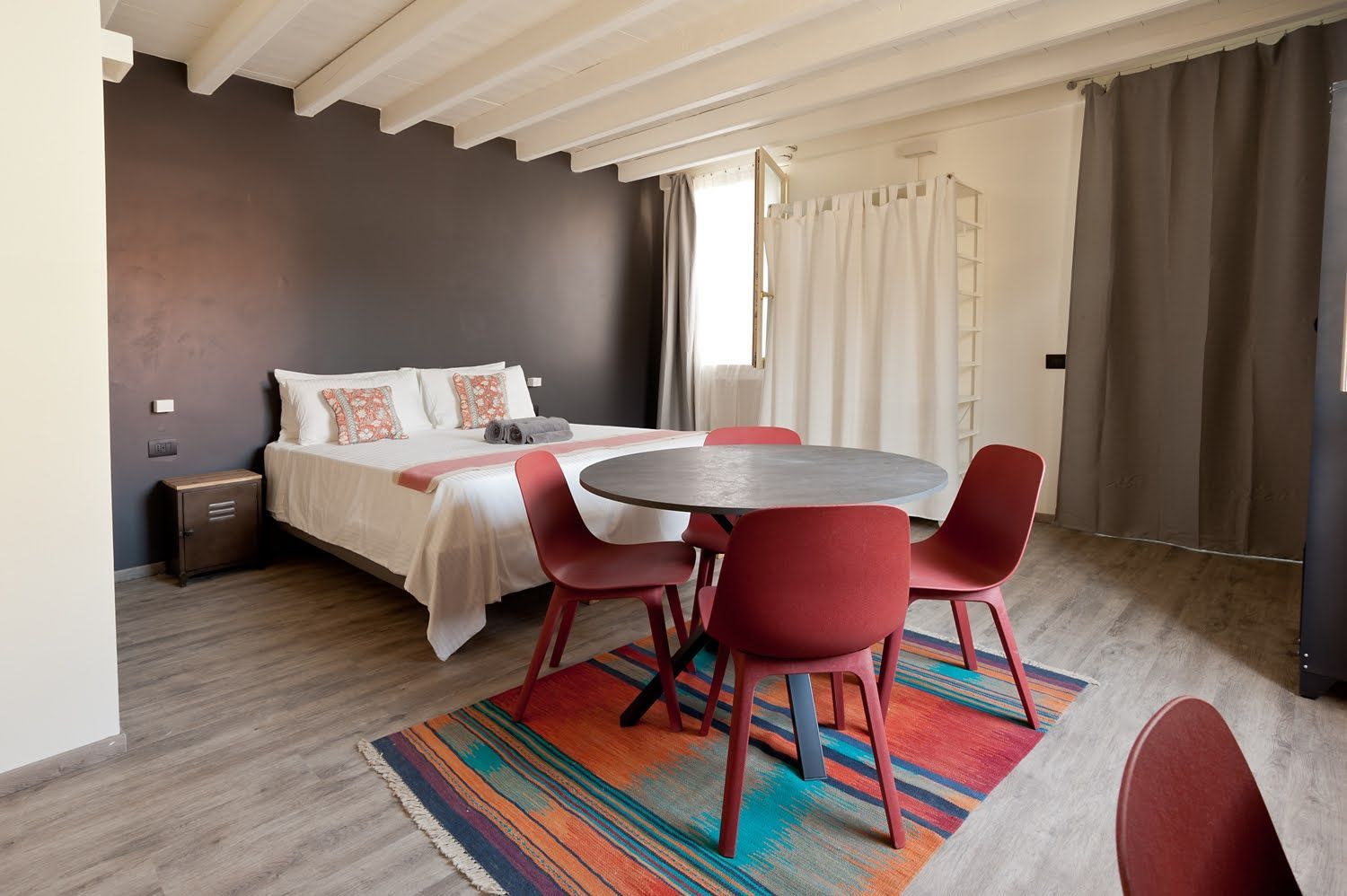 Camera da letto con letto, tavolo rotondo con sedie rosse e tappeto. Parete grigio scuro, soffitto bianco e tende.