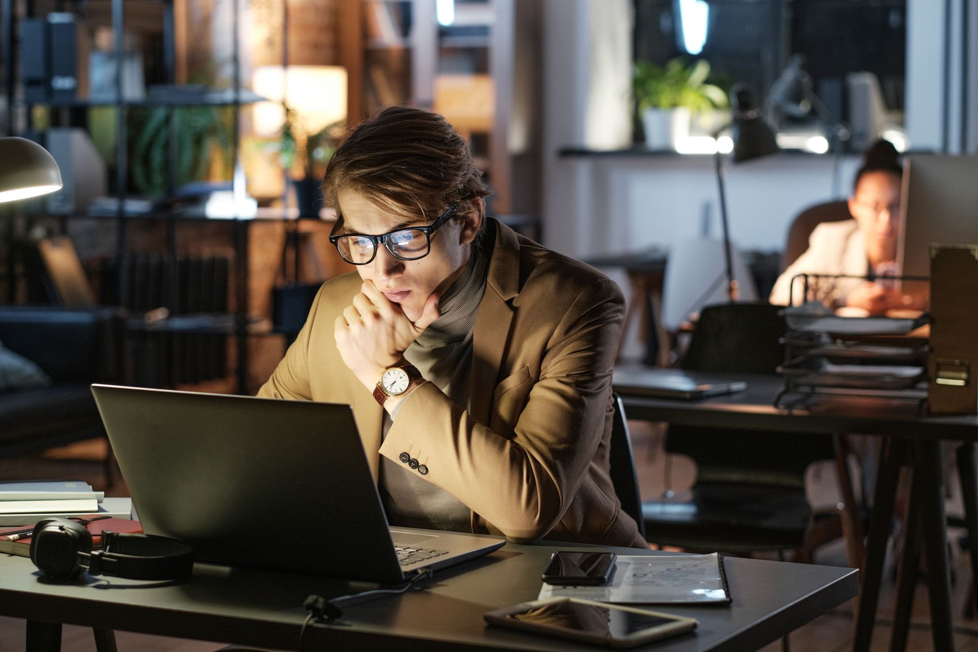 Uomo con gli occhiali che guarda scioccato il suo computer portatile mentre lavora fino a tardi in ufficio.